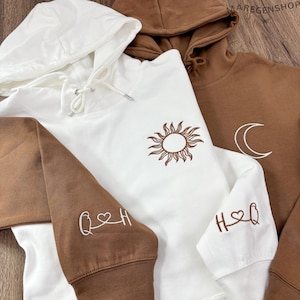 Könnte beinhalten: Drei Kapuzenpullover in Weiß und Braun. Der weiße Pullover hat ein Sonnendesign und braune Ärmel mit gestickten Initialen. Die braunen Sweatshirts haben ein Monddesign und Initialen. Die Sweatshirts haben Kordelzüge.