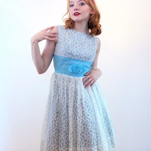 May include: A vintage-style sleeveless dress with a light blue lace overlay. The dress features a bright blue waist sash with a matching floral accent. The model is wearing red heels.