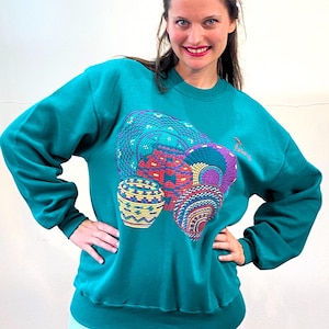 May include: Teal blue sweatshirt with a colorful embroidered design of woven baskets. The sweatshirt has a crew neck and long sleeves.