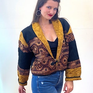 Peut inclure: Une veste à motifs noirs et bruns avec des bordures dorées et une fermeture à bouton. La veste présente un motif géométrique et est portée sur un haut noir et un jean bleu.