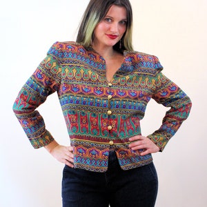 Pu&ograve; includere: Una blusa a maniche lunghe con bottoni e un motivo geometrico colorato. La blusa ha un collo a V e bottoni dorati.