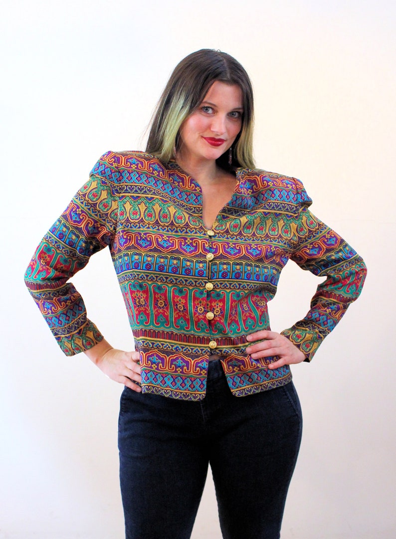 Pu&ograve; includere: Una blusa a maniche lunghe con bottoni e un motivo geometrico colorato. La blusa ha un collo a V e bottoni dorati.