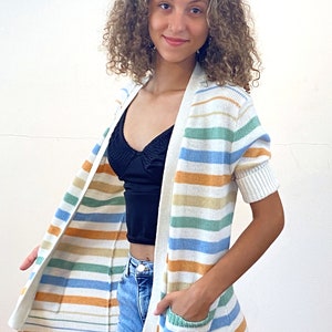 May include: A white striped cardigan with short sleeves and a pocket on the front. The cardigan has stripes of blue, green, orange, and beige.