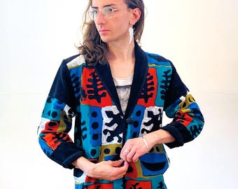 May include: A black velvet blazer with a colorful abstract print featuring geometric shapes, leaves, and dots in shades of red, blue, green, and white. The blazer has a button closure and long sleeves.
