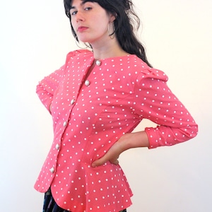 Könnte beinhalten: Eine rosa Blusen mit weißen Punkten, weißen Knöpfen und einem Peplum-Saum. Die Bluse hat lange Ärmel und einen runden Ausschnitt.
