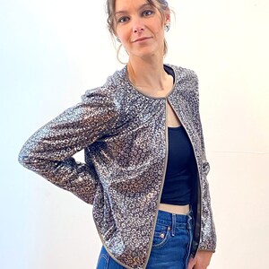 May include: A silver sequined jacket with a brown trim. The jacket has a round neckline and long sleeves. The model is wearing the jacket over a black tank top and blue jeans.
