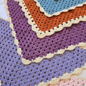May include: A collection of five triangular crochet bandanas arranged in a layered stack. Each piece features a classic granny square stitch pattern in solid colors of sage green, dusty blue, burnt orange, purple, and light pink, finished with a cream border.