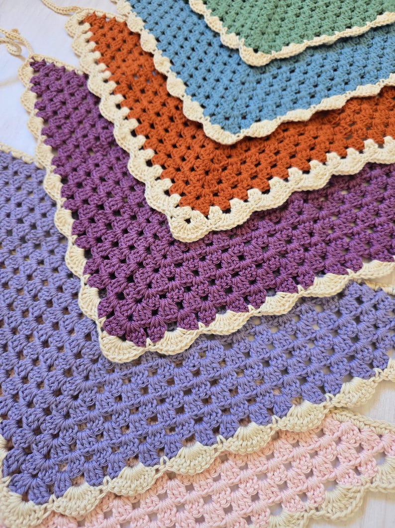 May include: A collection of five triangular crochet bandanas arranged in a layered stack. Each piece features a classic granny square stitch pattern in solid colors of sage green, dusty blue, burnt orange, purple, and light pink, finished with a cream border.