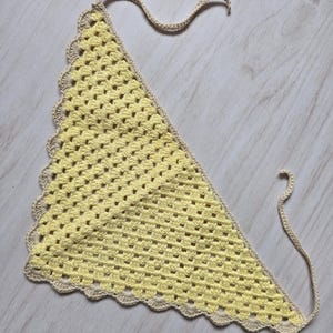 May include: A triangular crochet bandana featuring a pale yellow center with a textured granny stitch pattern. The edges are finished with a scalloped border in a light beige yarn, and two long matching beige ties extend from the top corners.