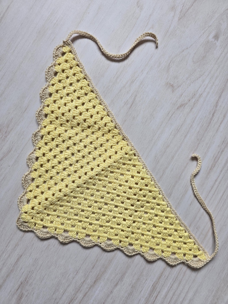 May include: A triangular crochet bandana featuring a pale yellow center with a textured granny stitch pattern. The edges are finished with a scalloped border in a light beige yarn, and two long matching beige ties extend from the top corners.