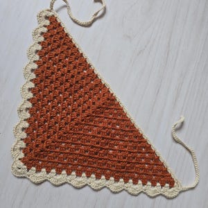 May include: A triangular crochet bandana featuring a rust-colored center with a classic granny square stitch pattern. The edges are finished with a cream-colored scalloped border and two long ties at the top corners for securing the garment.