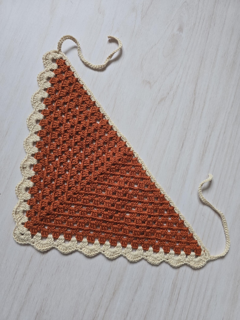May include: A triangular crochet bandana featuring a rust-colored center with a classic granny square stitch pattern. The edges are finished with a cream-colored scalloped border and two long ties at the top corners for securing the garment.