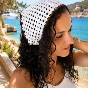 May include: A crochet headscarf featuring a white openwork granny square pattern with a solid dark brown border. The triangular head covering is constructed from yarn and displays a distinct geometric texture throughout the fabric design.