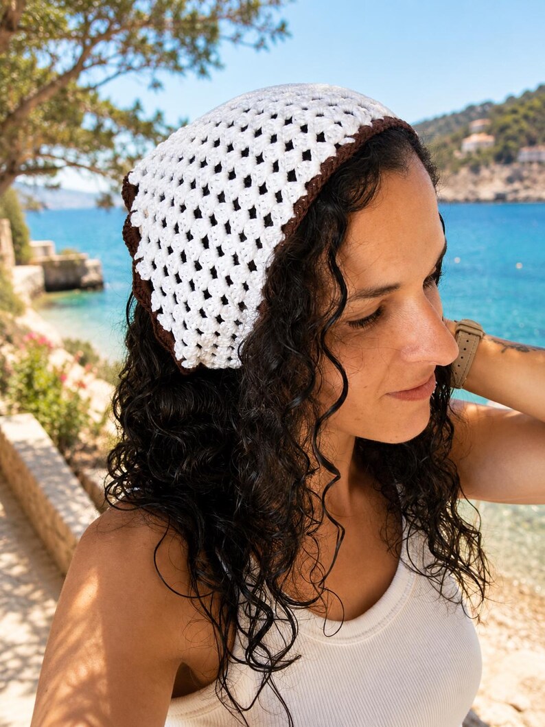May include: A crochet headscarf featuring a white openwork granny square pattern with a solid dark brown border. The triangular head covering is constructed from yarn and displays a distinct geometric texture throughout the fabric design.