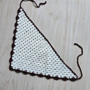 May include: A triangular crochet bandana featuring a white granny square pattern with a dark brown scalloped border and two thin brown ties at the top corners. The accessory is crafted with a repeating open-work stitch design throughout the center.