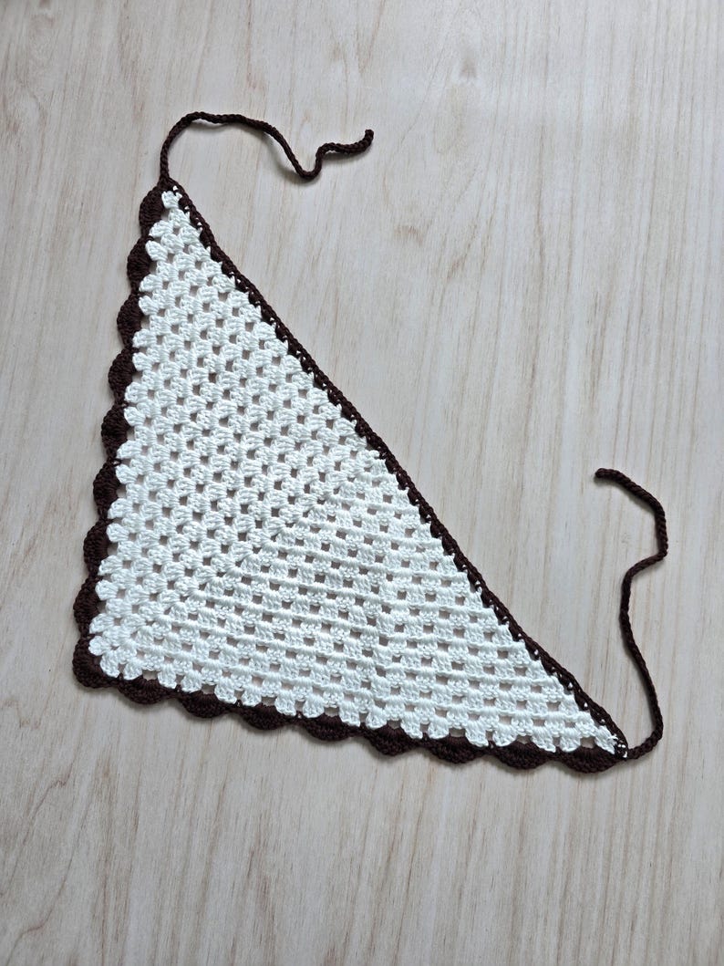 May include: A triangular crochet bandana featuring a white granny square pattern with a dark brown scalloped border and two thin brown ties at the top corners. The accessory is crafted with a repeating open-work stitch design throughout the center.