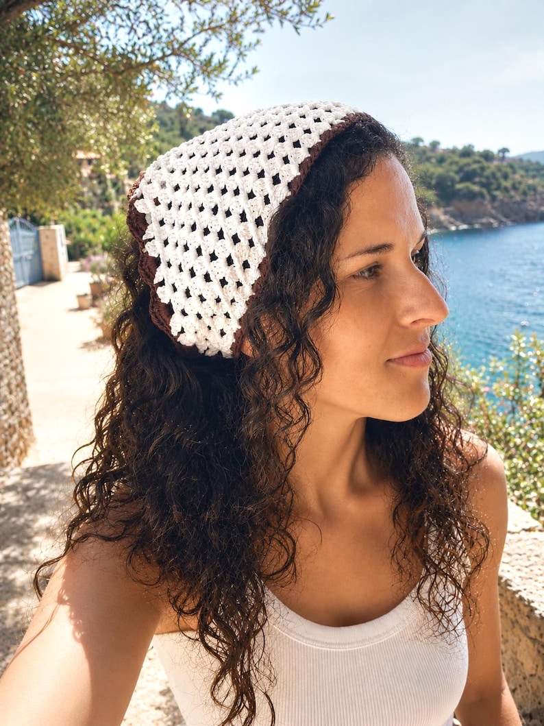 May include: A triangular crochet headscarf featuring a white openwork diamond pattern with a solid dark brown border. The headscarf is constructed with a textured yarn and is tied at the back of the head, covering the crown with a classic triangular shape.