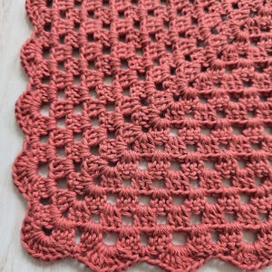 May include: A dusty rose crochet doily featuring a square grid pattern made of interconnected yarn stitches. The edges of the textile are finished with a decorative scalloped border consisting of rounded loops that create a wavy perimeter.