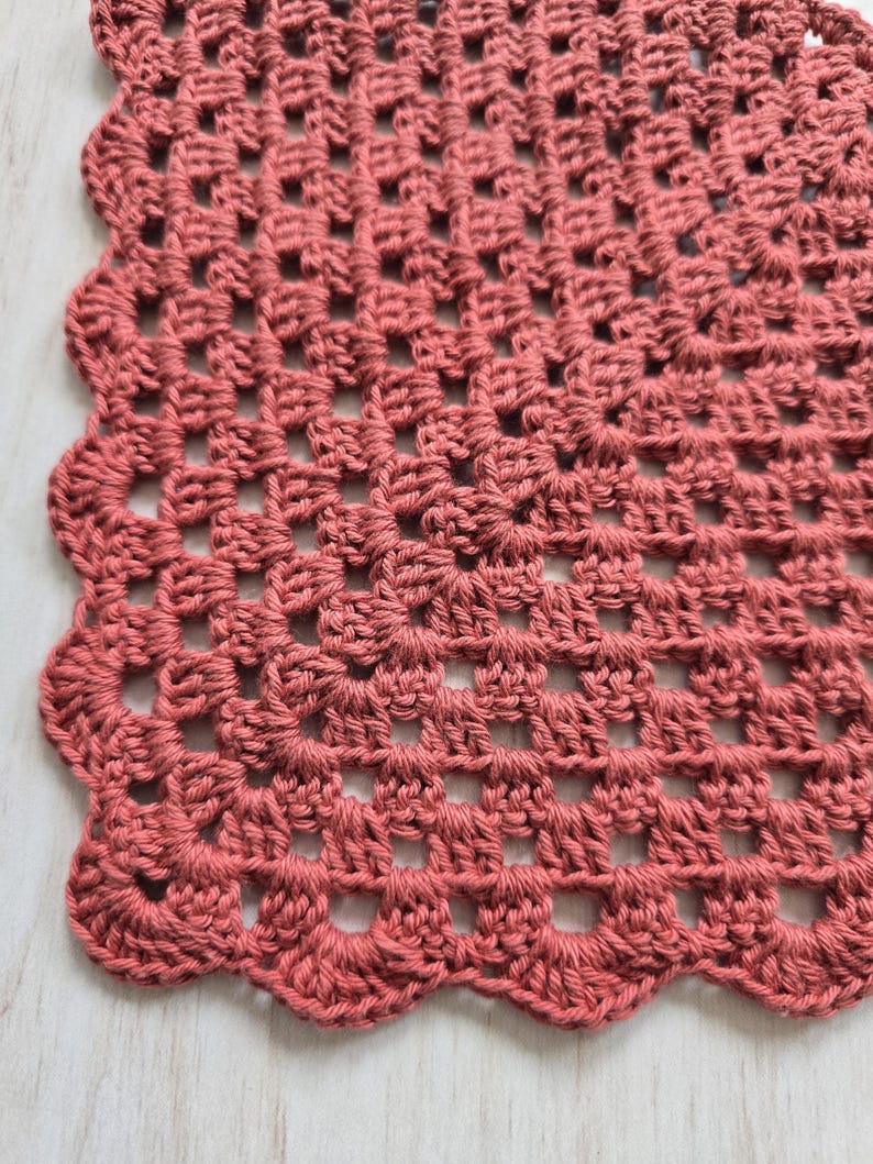 May include: A dusty rose crochet doily featuring a square grid pattern made of interconnected yarn stitches. The edges of the textile are finished with a decorative scalloped border consisting of rounded loops that create a wavy perimeter.