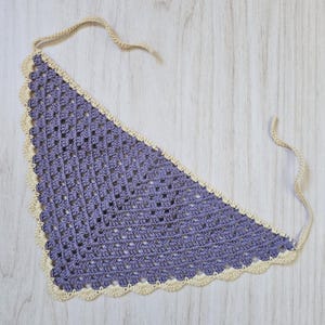 May include: A triangular crochet bandana featuring a purple granny square pattern with a cream-colored scalloped border and two thin matching cream-colored ties extending from the top corners for securing the garment around the neck.