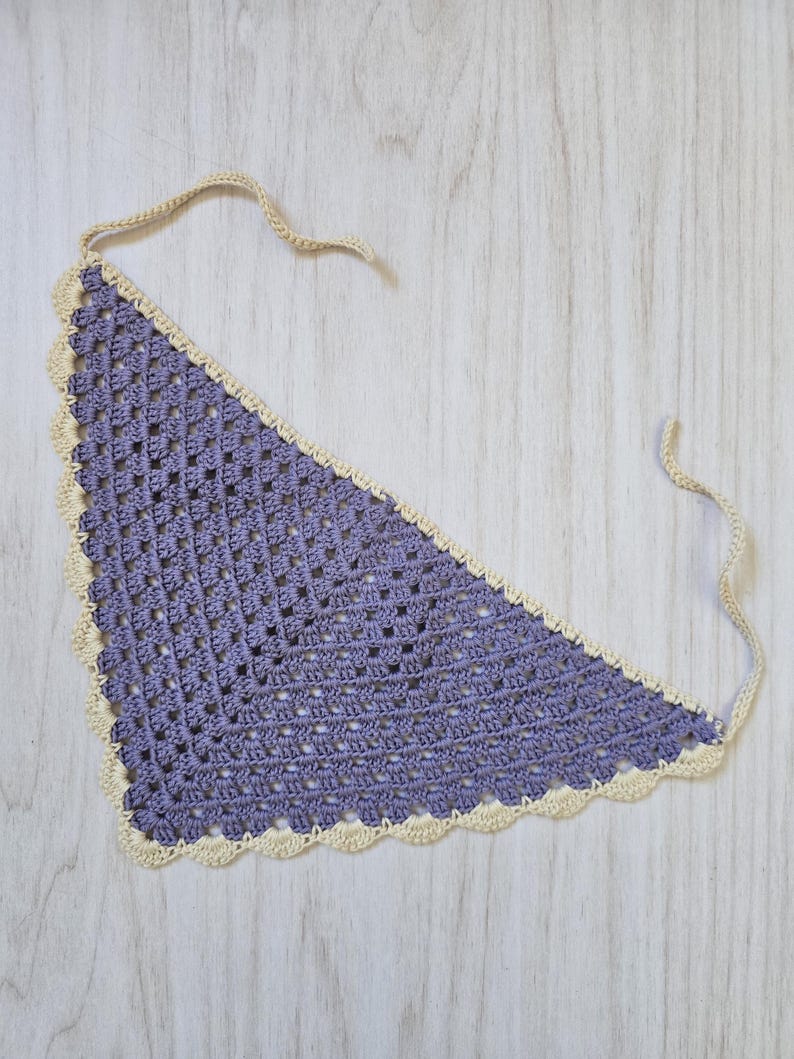 May include: A triangular crochet bandana featuring a purple granny square pattern with a cream-colored scalloped border and two thin matching cream-colored ties extending from the top corners for securing the garment around the neck.