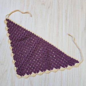 May include: A triangular crochet bandana featuring a deep purple center with a textured, open-stitch pattern. The edges are finished with a scalloped cream-colored border and include two long, thin cream-colored ties at the top corners for fastening.