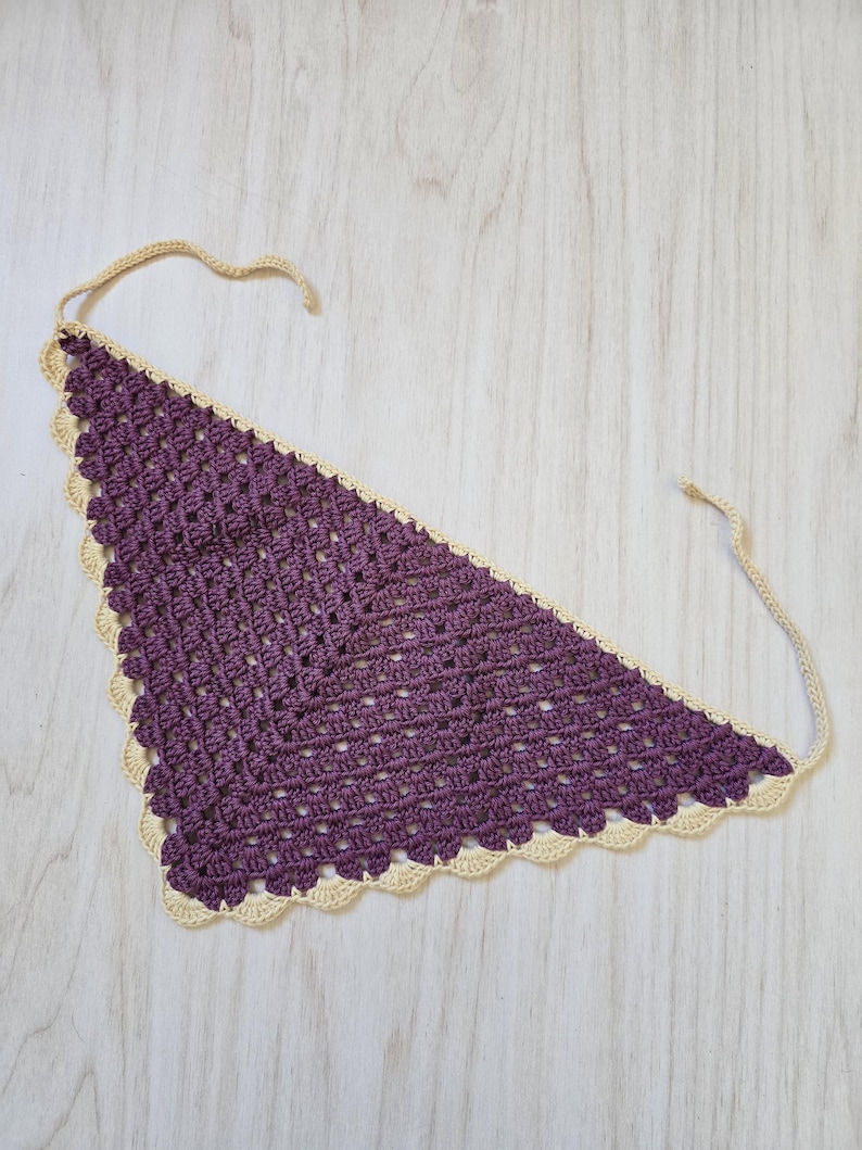 May include: A triangular crochet bandana featuring a deep purple center with a textured, open-stitch pattern. The edges are finished with a scalloped cream-colored border and include two long, thin cream-colored ties at the top corners for fastening.