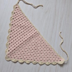 May include: A triangular crochet bandana featuring a light pink granny square pattern. The accessory is bordered with a scalloped edge in a cream color and includes two thin crocheted ties at the top corners for securing the garment in place.