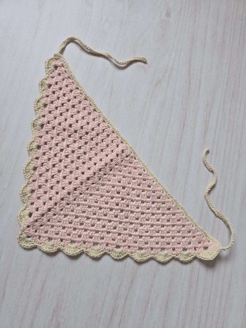 May include: A triangular crochet bandana featuring a light pink granny square pattern. The accessory is bordered with a scalloped edge in a cream color and includes two thin crocheted ties at the top corners for securing the garment in place.