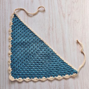 May include: A triangular crochet bandana featuring a deep blue center with a repeating square stitch pattern. The edges are finished with a scalloped border in a light cream color, and two thin matching cream ties extend from the top corners.