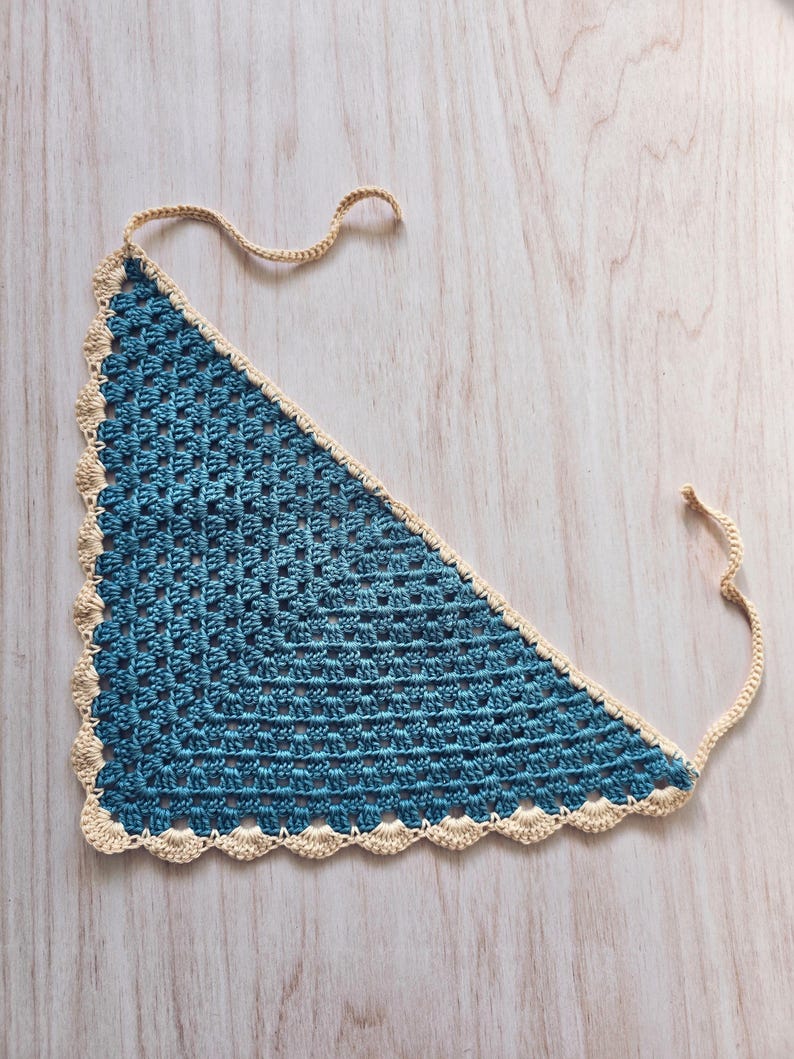 May include: A triangular crochet bandana featuring a deep blue center with a repeating square stitch pattern. The edges are finished with a scalloped border in a light cream color, and two thin matching cream ties extend from the top corners.