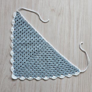 May include: A triangular crochet bandana featuring a light blue granny square pattern with a white scalloped border and two thin white ties extending from the top corners, crafted with a consistent open-stitch texture throughout the main body.