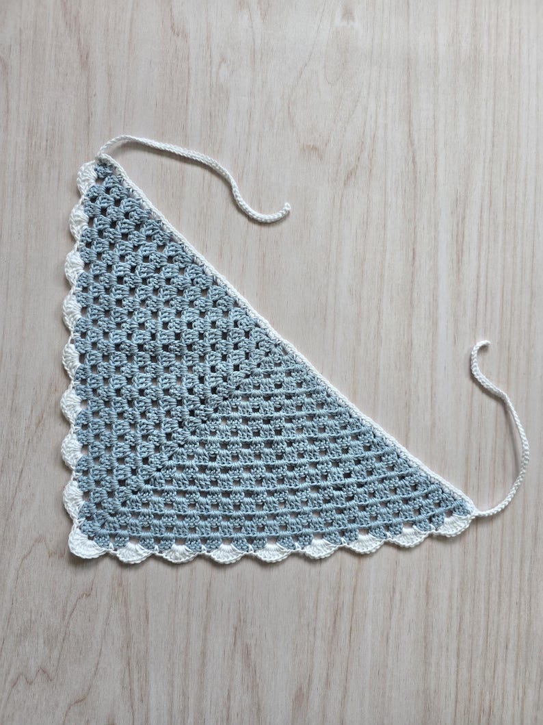 May include: A triangular crochet bandana featuring a light blue granny square pattern with a white scalloped border and two thin white ties extending from the top corners, crafted with a consistent open-stitch texture throughout the main body.