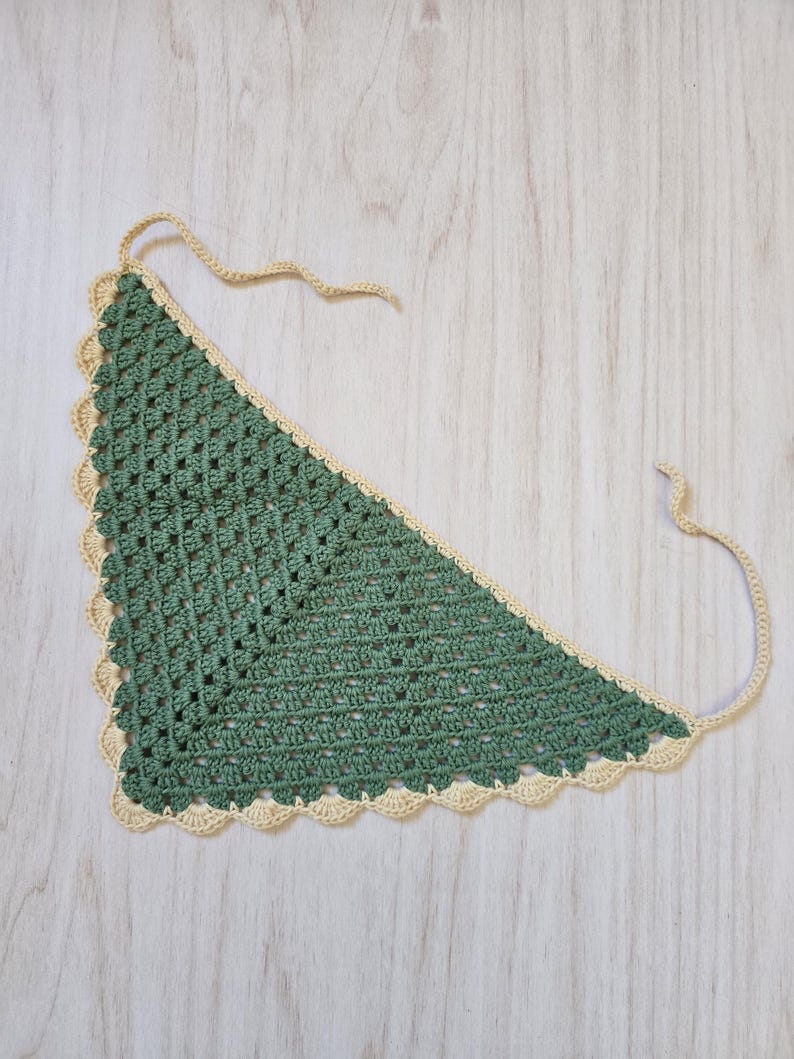 May include: A triangular crochet bandana featuring a sage green center with a textured openwork pattern and a scalloped cream-colored border. The accessory includes two thin crocheted ties at the top corners for securing the garment in place.