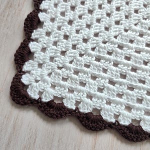May include: A white crocheted doily featuring a square grid pattern made of yarn. The edges of the textile are finished with a decorative scalloped border in a dark brown color, creating a distinct contrast against the white center section.