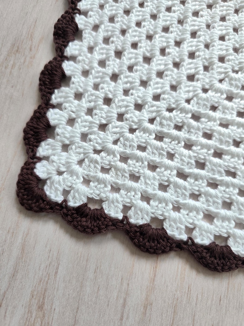 May include: A white crocheted doily featuring a square grid pattern made of yarn. The edges of the textile are finished with a decorative scalloped border in a dark brown color, creating a distinct contrast against the white center section.