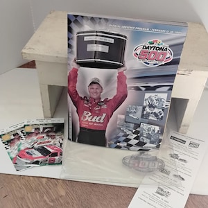 Può includere: Programma ricordo della Daytona 500 del 12-20 febbraio 2005, con un pilota che tiene un trofeo. Include un'immagine di auto da corsa e un motivo a scacchiera. Include anche un biglietto con il logo Daytona 500.