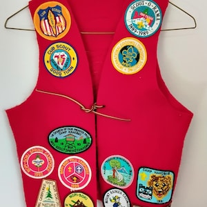 May include: A red Cub Scout vest with multiple patches, including "Cub Scout," "Scout-O-Rama," and camp names. The vest has a leather cord closure and is displayed on a hanger. Patches feature various colors and designs.