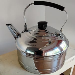 May include: Shiny, stainless steel tea kettle with a black handle and lid knob. The kettle has a spout and a curved handle. It sits on a small, white wooden stool. The kettle is reflective, showing the surrounding environment.
