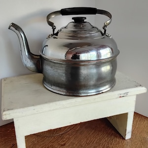 Może przedstawiać: Stalowy czajnik z czarnym uchwytem i wylewką. Czajnik stoi na małym, białym, drewnianym stołku. Czajnik ma błyszczącą, odblaskową powierzchnię, a stołek ma postarzany wygląd. Artykuł kuchenny.