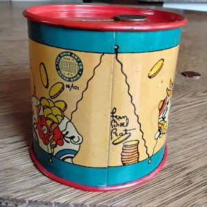 Könnte beinhalten: Eine antike Metall-Sparbüchse mit einer Cartoon-Illustration eines Schweins, das einen roten Hut trägt und eine Münze hält. Die Sparbüchse ist in leuchtenden Farben bemalt, darunter Gelb, Rot, Blau und Grün. Die Sparbüchse hat einen roten Deckel mit einem Schlitz für Münzen. Die Sparbüchse trägt ein Etikett mit der Aufschrift "The Ohio Art Co. Bryan, Ohio 14/101".
