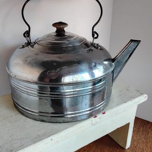 May include: A vintage, shiny metal kettle with a black handle and spout. The kettle has a wooden knob on the lid and sits on a small, white, distressed wooden bench. The kettle is a classic kitchenware item.