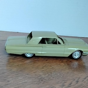 Puede incluir: Un modelo de coche de color beige con techo rígido. El coche tiene neumáticos de banda blanca y tapacubos cromados. El modelo está sobre una superficie de madera.