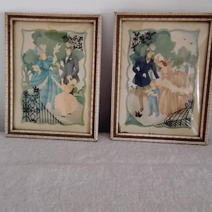 May include: Two framed vintage prints depicting scenes of people in a garden setting. The prints feature figures in period clothing, with a color palette of blues, pinks, and greens. The frames are rectangular with a cream and gold trim.