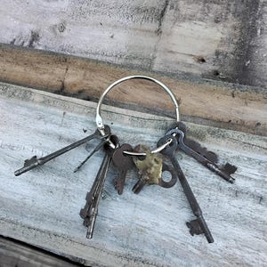 May include: A set of antique keys attached to a silver key ring. The keys are of different sizes and designs, with a variety of metal finishes, including brass and iron. The keys are placed on a weathered wooden surface.