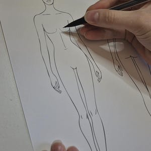 Puede incluir: Ilustración de moda en progreso, con una figura calva dibujada con tinta negra sobre papel blanco. Una mano sostiene un bolígrafo negro, agregando detalles al torso de la figura. En el fondo se ve otro contorno de una figura.