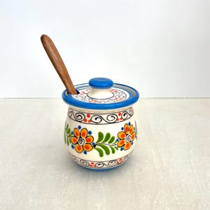May include: A ceramic jar with a lid and wooden spoon. The jar is white with a blue rim and lid, and is decorated with orange, yellow, and green floral designs. The spoon is brown.