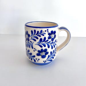 May include: A ceramic mug with a cream-colored base featuring a hand-painted cobalt blue floral and leafy vine pattern. The mug has a rounded body, a sturdy handle, and a solid blue rim, showcasing a repeating botanical design.