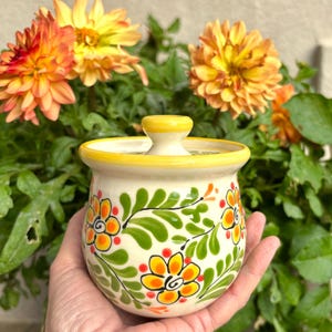 May include: A hand-painted ceramic jar with a lid, featuring a cream-colored base adorned with orange and yellow floral designs and green leaves. The rim and lid handle are yellow. The jar is held in front of a backdrop of orange and yellow flowers.