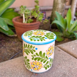 May include: A cylindrical ceramic jar with a matching lid, featuring a cream-colored base decorated with hand-painted yellow flowers, green fern-like leaves, and small blue and orange dots, finished with a bright blue rim.